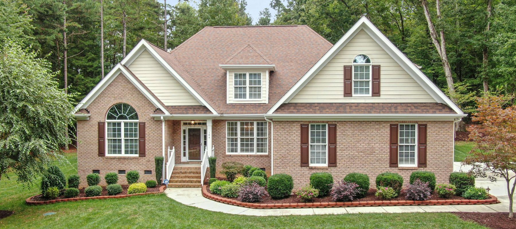 A two-story suburban brick house with neatly trimmed shrubs and a backdrop of tall trees