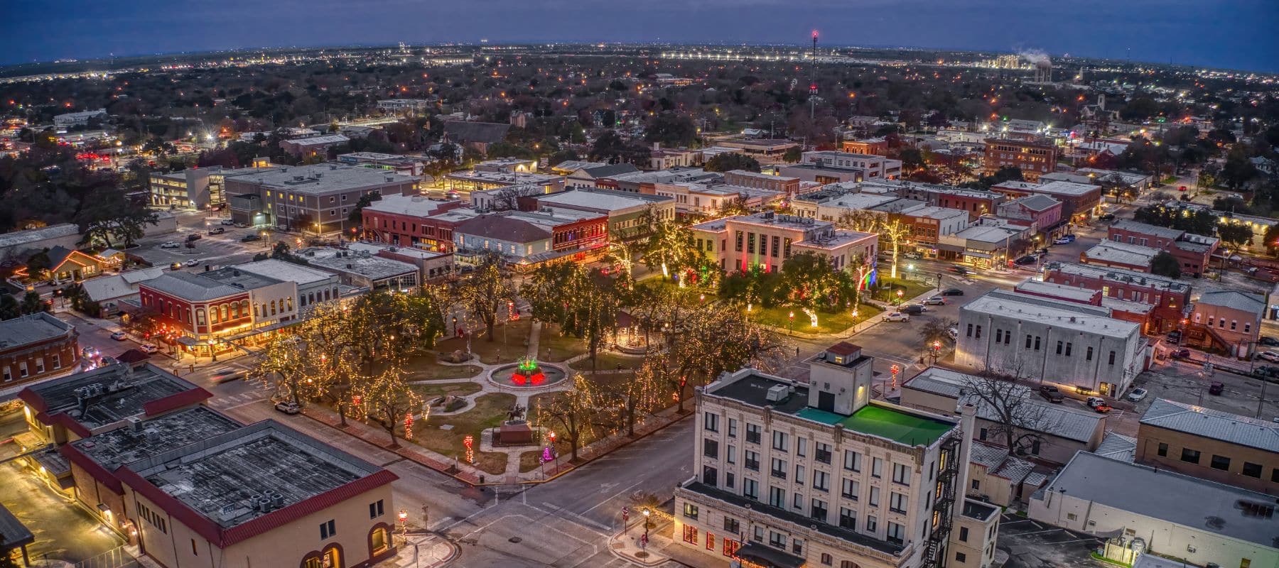Aerial view of a historic downtown area lit up at dusk, featuring a central park with festive lights and surrounding buildings under a dramatic evening sky